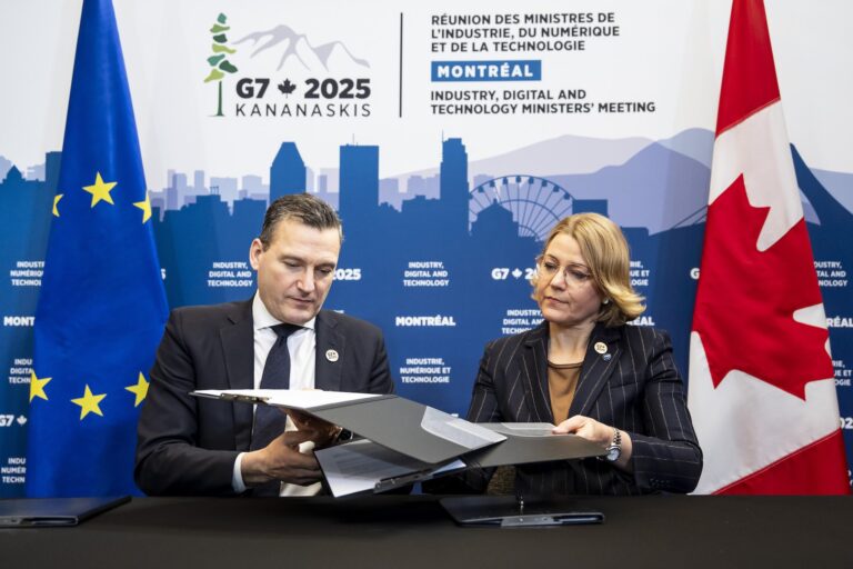 A shot of Evan Solomon and Henna Virkkunen trading document during a signing photo-op, against a backdrop featuring the G7 2025 logo. There are Canadian and EU flags in the background.