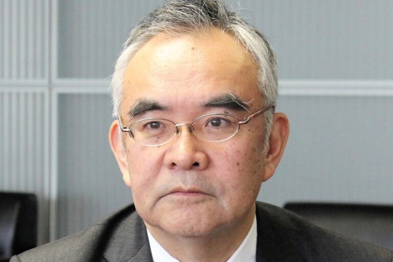 A headshot of Kanji Yamanouchi in a meeting room setting. He is wearing a suit and small, half-frame glasses