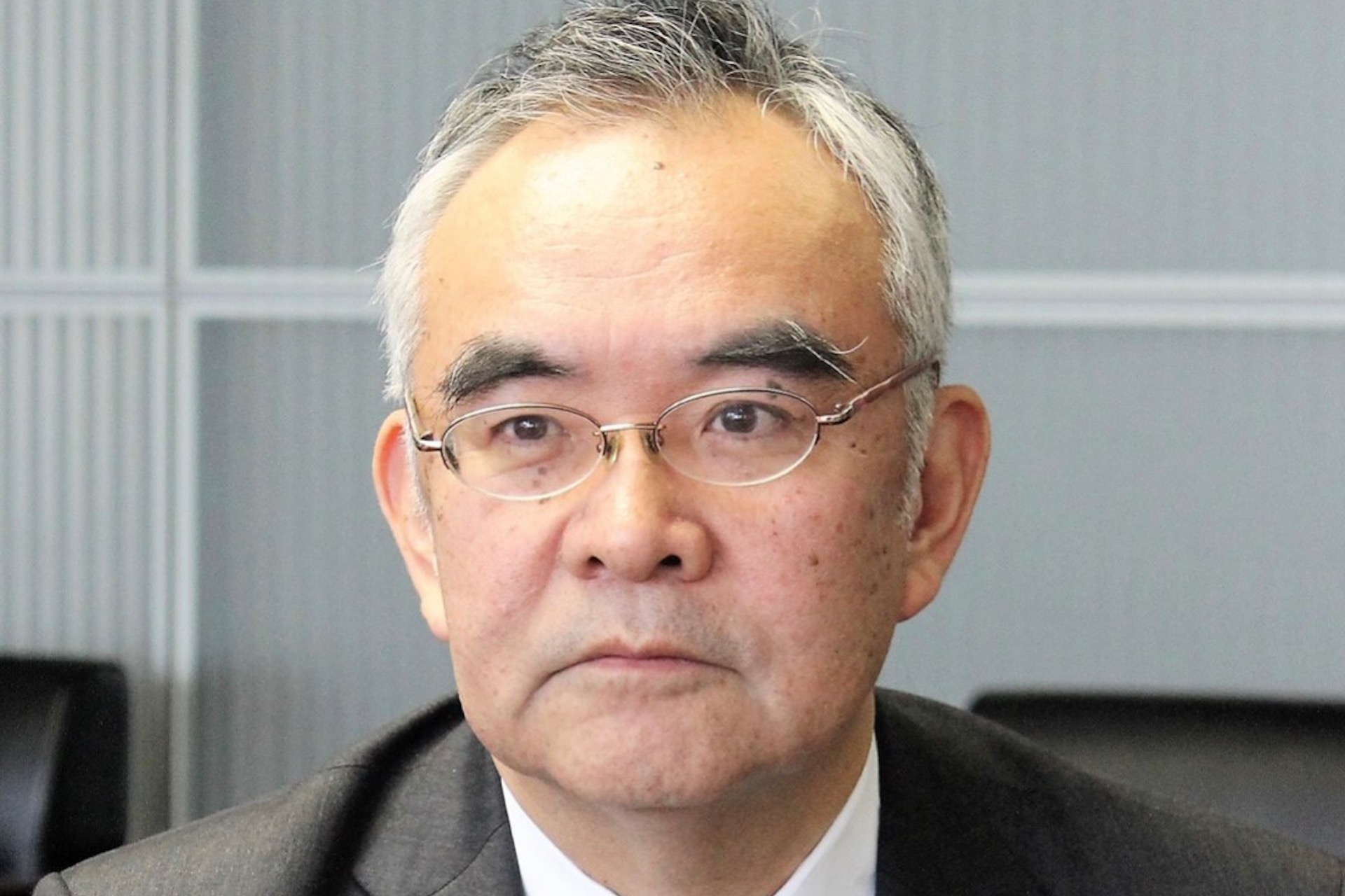 A headshot of Kanji Yamanouchi in a meeting room setting. He is wearing a suit and small, half-frame glasses