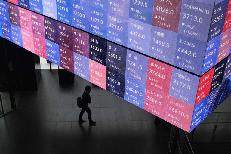 View from above of a large monitor hanging from the ceiling split into dozens of stock market ticker quotes. A shadowed figure walks past below the board.