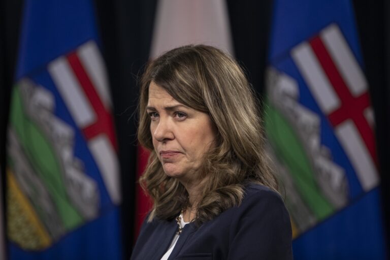 Danielle Smith appears in front of blurred Alberta flags, looking serious.