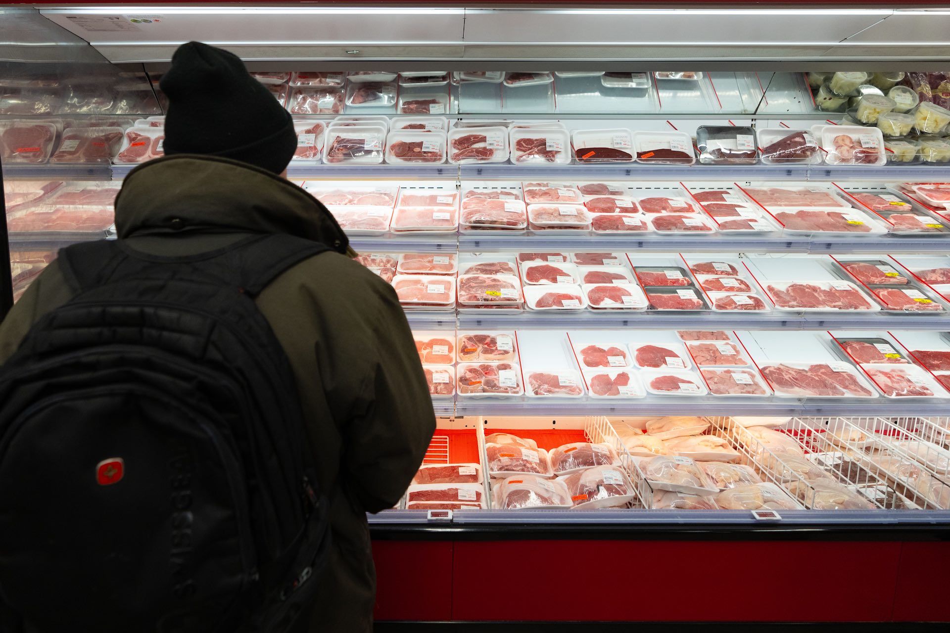 A shot from behind of a shopper in winter clothing looking at the meat shelves in a supermarket.