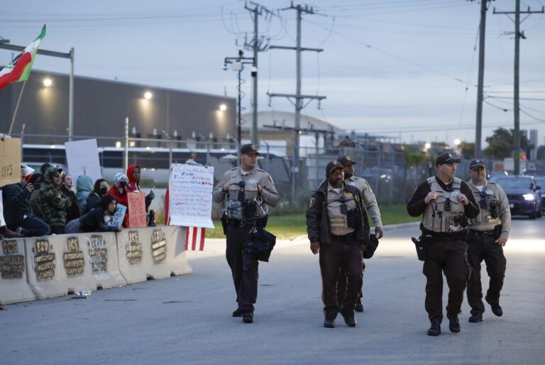 A street-level photo of five police officers walking toward the camera, with a cluster of protesters behind a concrete barrier on the left of the frame. There is a large building behind chainlink fencing in the background.