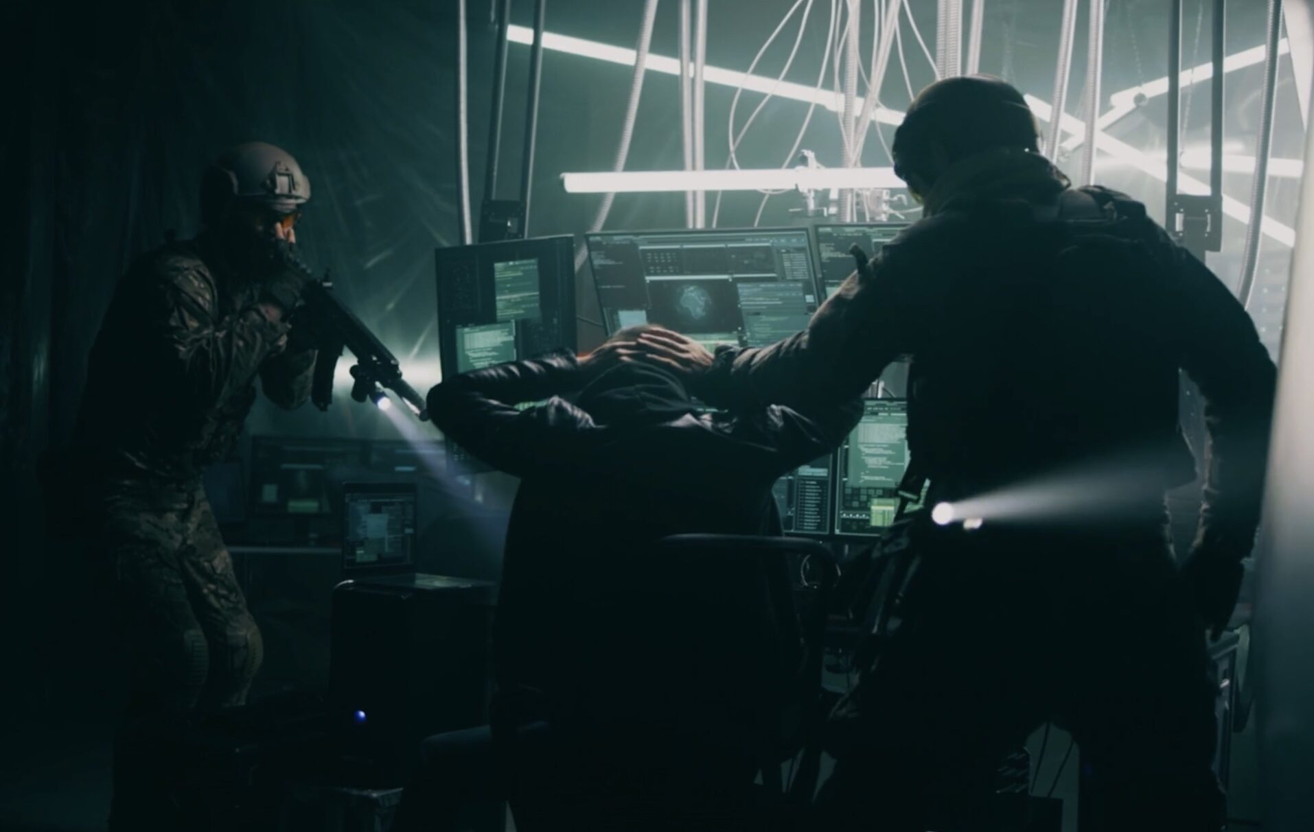 A screen capture from a video showing officers in tactical combat gear arresting a man, who is seated before an array of computer screens in a dark setting.