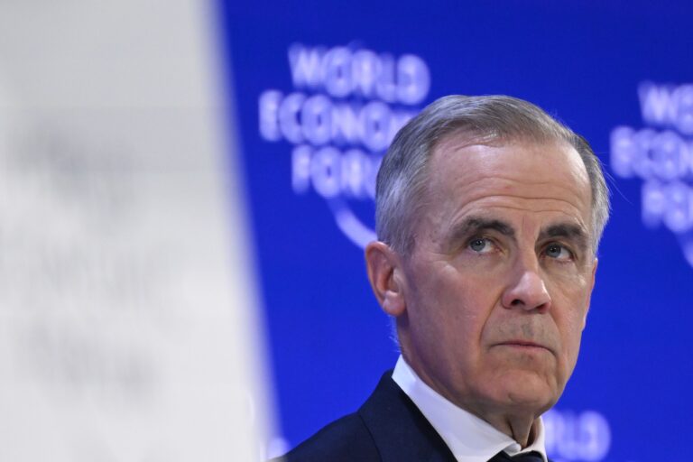 A close-up of Mark Carney wearing a solemn expression, with the World Economic Forum logo on a blue backdrop behind him.