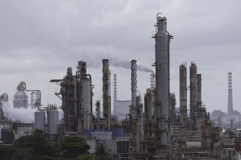 A wide shot of the stacks and distilling towers of an oil refinery, against a flat grey sky.