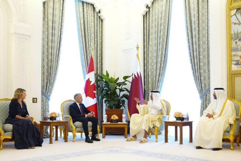 A wide shot of Mark Carney and Melanie Joly seated in an ornate room with high ceilings, conversing with the Emir of Qatar and another person. There are Canadian and Qatari flags in the corner between them.