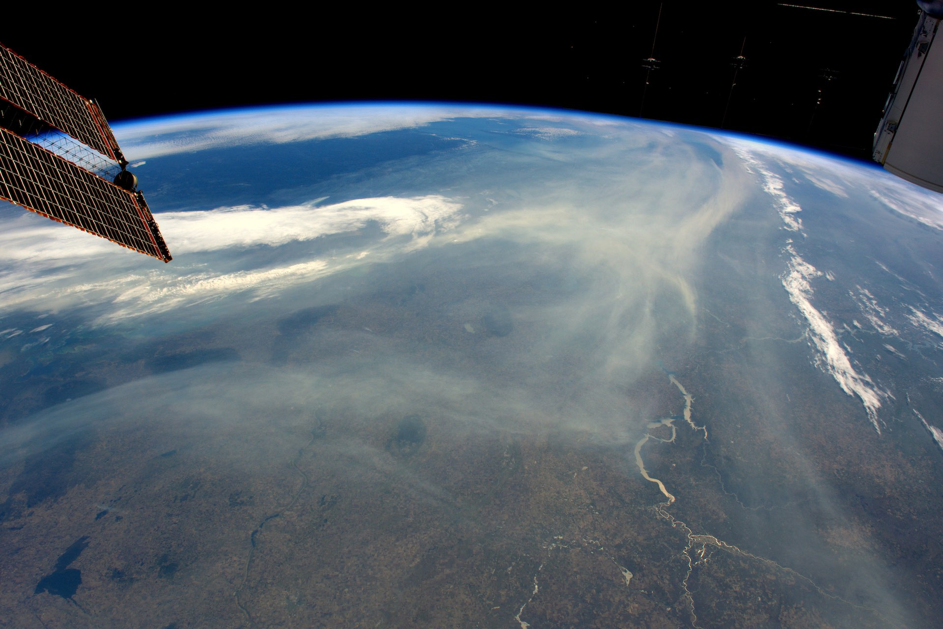 A photo from the International Space Station of northwestern Canada, occluded in part by trails of wildfire smoke. One of the station's solar arrays is visible in the top left corner of the shot.