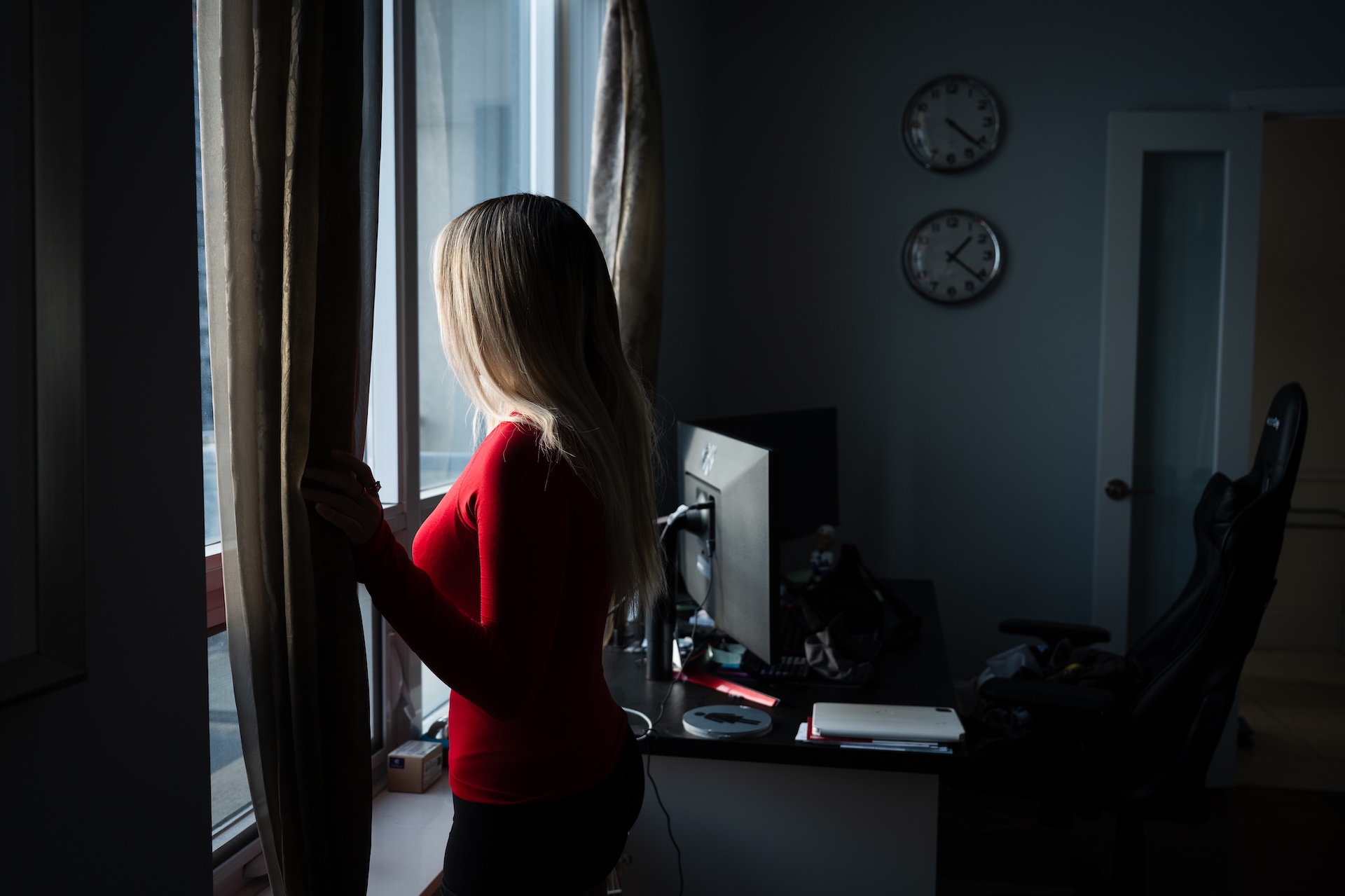 A girl in a red sweater looks thoughtfully out a window in a dimly-lit room. A desk with a computer and clocks on the wall are visible; a calm atmosphere prevails.