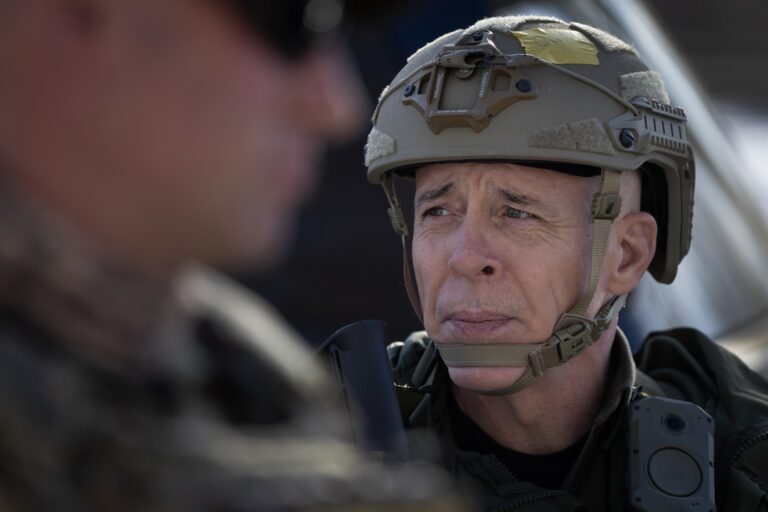 Image of Greg Bovino in tactical gear, looking to his right. Another person wearing dark glasses can be seen out-of-focus in the extreme foreground on the left side of the picture.