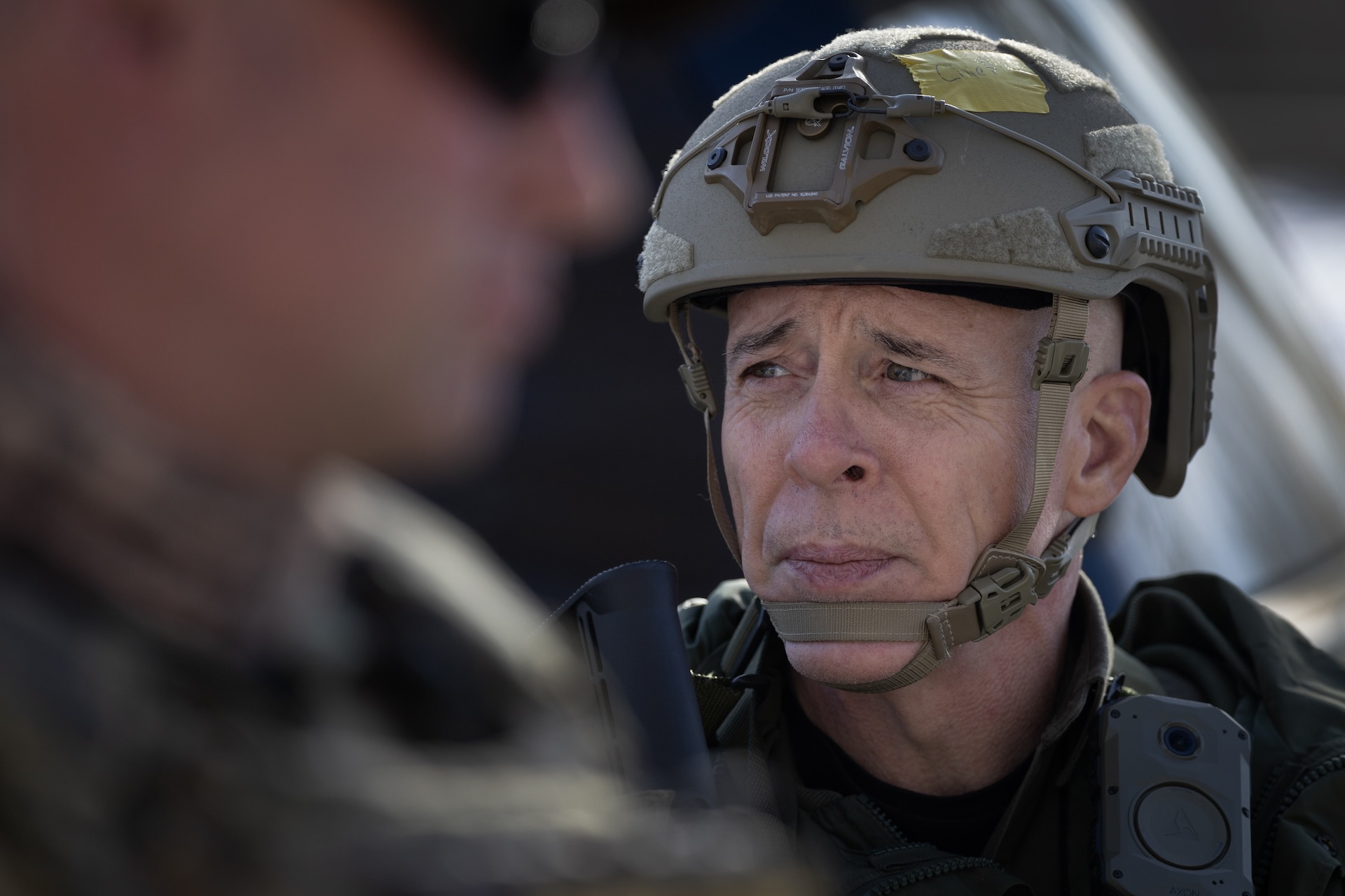Image of Greg Bovino in tactical gear, looking to his right. Another person wearing dark glasses can be seen out-of-focus in the extreme foreground on the left side of the picture.