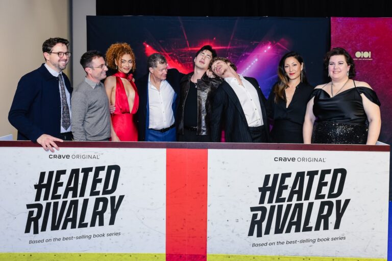 A cast shot of producers and actors from Heated Rivalry, behind a set of fake hockey arena boards. The actors who play the two principal characters are doing mock swoons for each other.