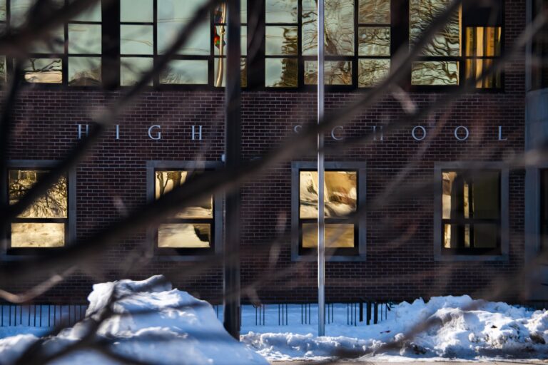 Brick high school in winter, with snow on the ground. Bare tree branches in the foreground. Reflective windows create a serene tone.