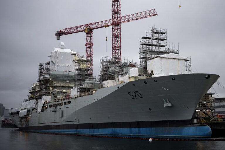 A shot of a joint support naval ship at the dock, but still under construction. There is scaffolding on its decks and cranes looming above it.