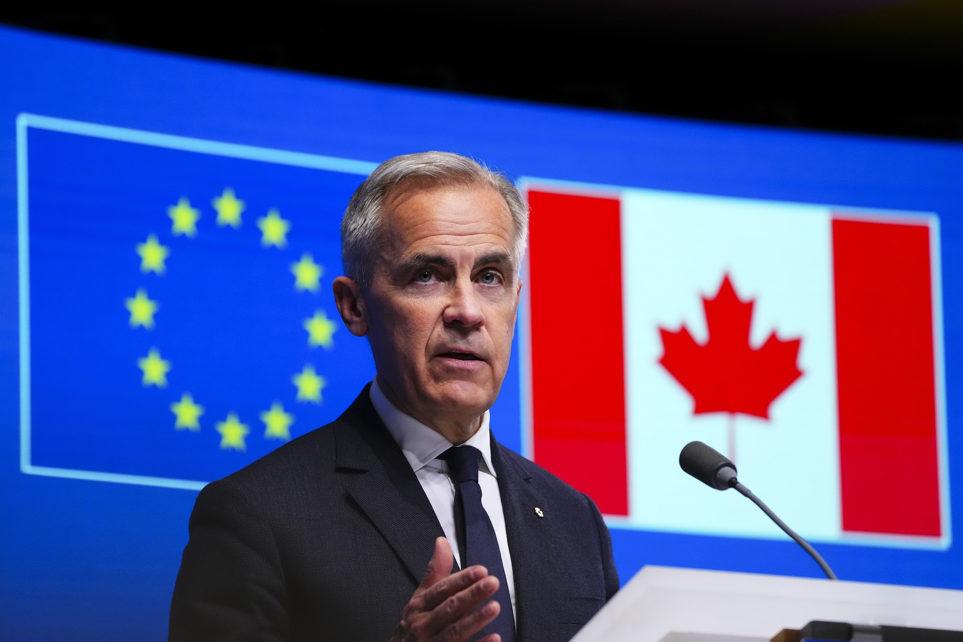 Canadian Prime Minister Mark Carney in a suit speaks at a podium with the EU and Canadian flags displayed on a screen behind him.