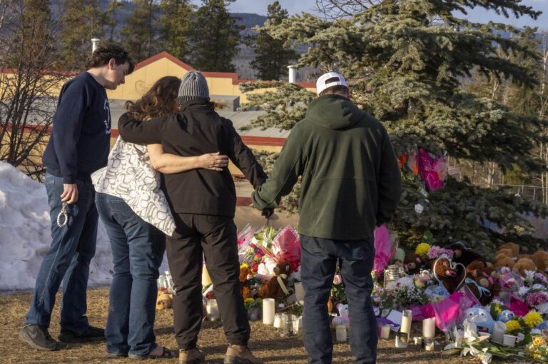 A photo from behind of four people viewing a cluster of flowers, stuffed animals and candles on the ground beneath an evergreen tree. Two have arms around each other. The Tumbler Ridge high school is in the background.