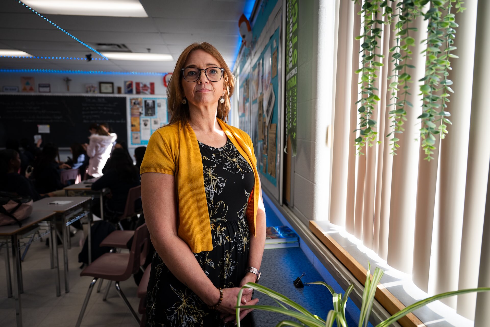 A woman in a bright yellow cardigan stands confidently in a classroom, with students and desks in the background. Soft light filters through blinds, creating a warm atmosphere.