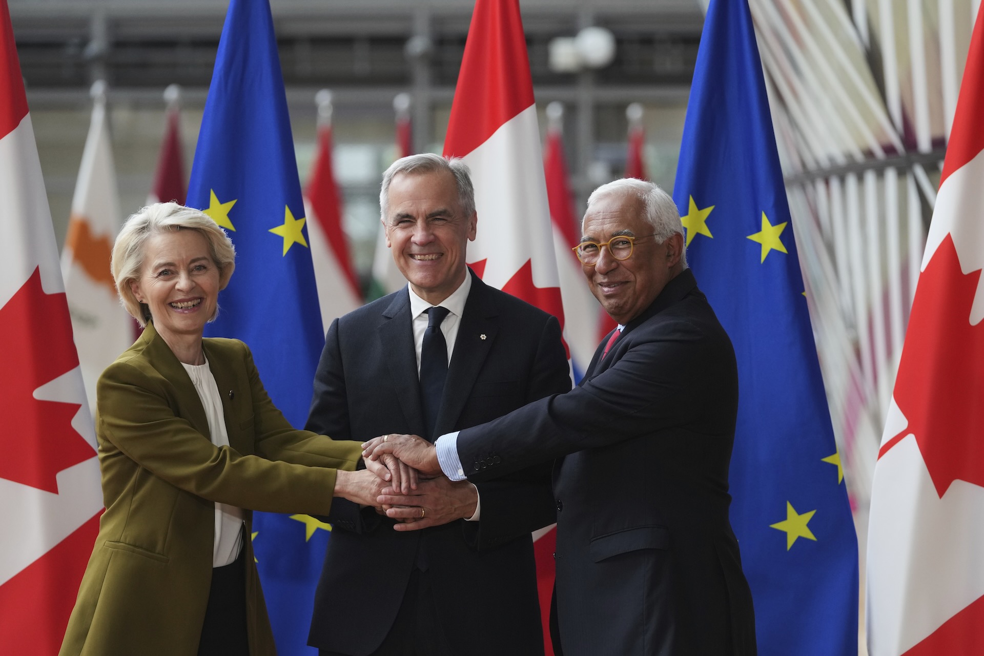 European Commission President Ursula von der Leyen, left; Canadian Prime Minister Mark Carney, centre and European Council President António Costa are smiling, holding hands in a unity gesture, standing before Canadian and EU flags, conveying co-operation and friendship.