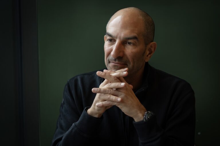 Andrew Lutfy sits against a green background, wearing a dark sweater and a watch. He holds his hands clasped in front of him and looks slightly away from the camera.