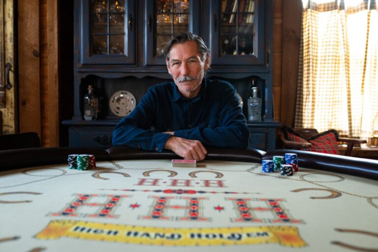 A portrait of Barry Dusome seated at a poker table with chips and a deck of cards in front of him. The setting is a wood-panelled hobby room decorated with reclaimed pub signs and rustic furniture.