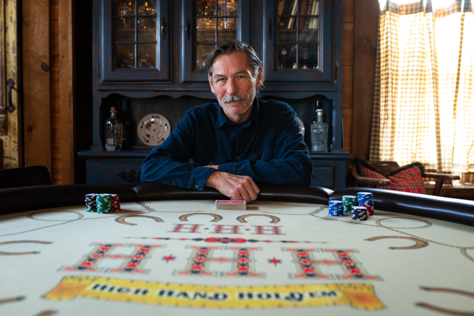 A portrait of Barry Dusome seated at a poker table with chips and a deck of cards in front of him. The setting is a wood-panelled hobby room decorated with reclaimed pub signs and rustic furniture.