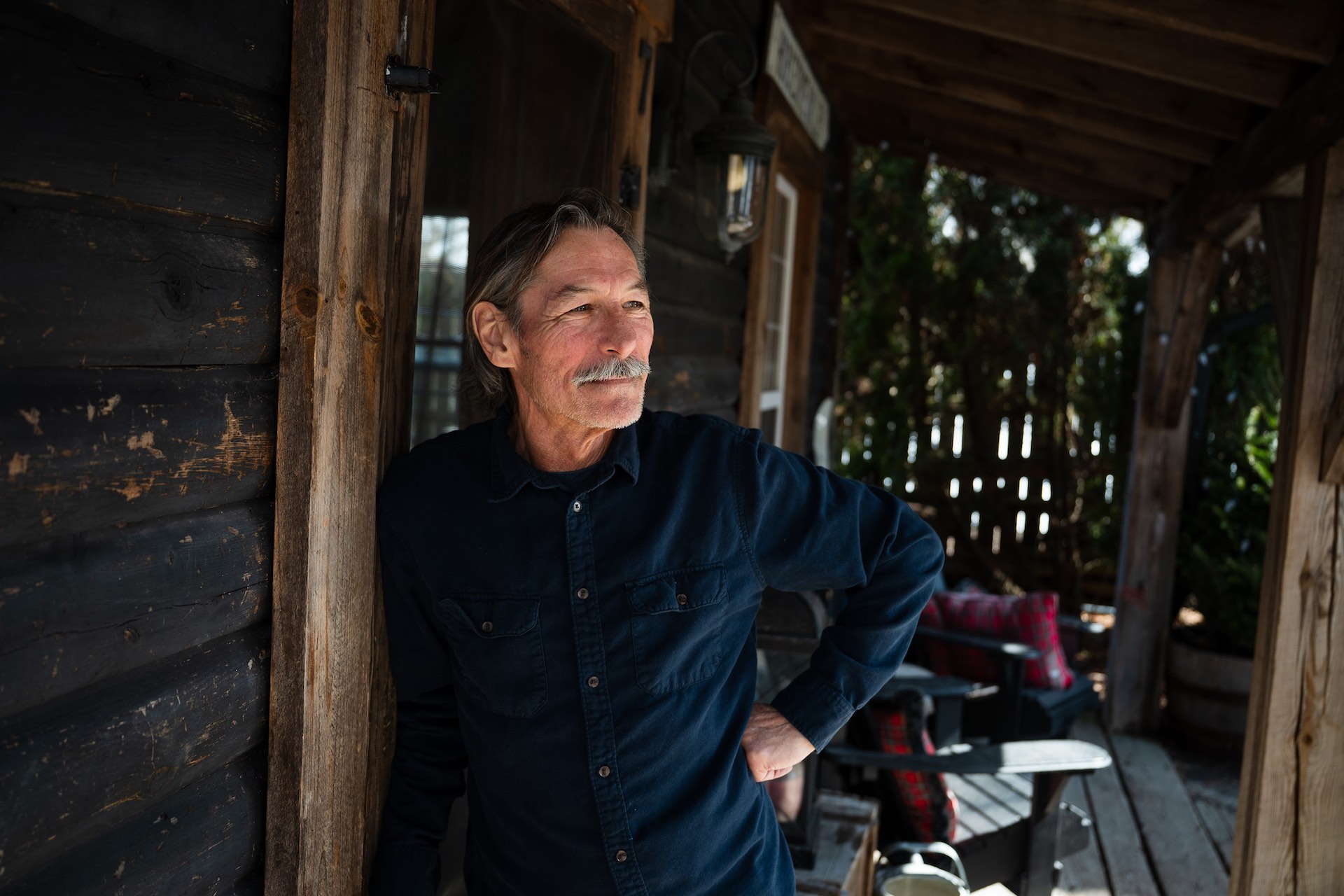 A portrait of Barry Dusome leaning against the door jamb of a rustic-looking porch. There are two Muskoka chairs in the background.