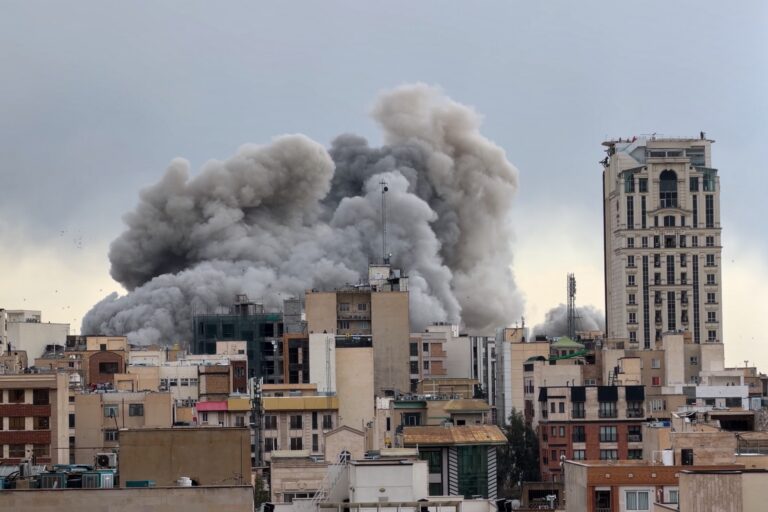 An image taken across rooftops in Tehran of a plume of dust and smoke from a bomb falling. There are midrise residential buildings in the foreground.