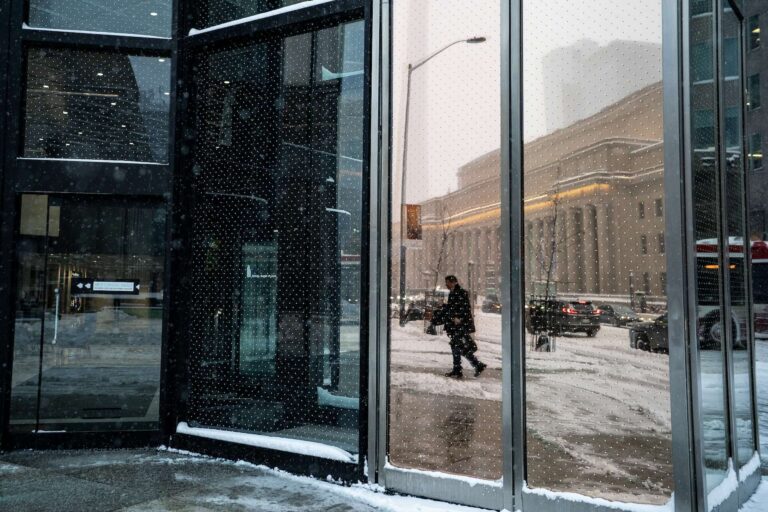 A commuter in a black coat is seen reflected in a glass door, walking along a snow-covered street with cars driving by in the background