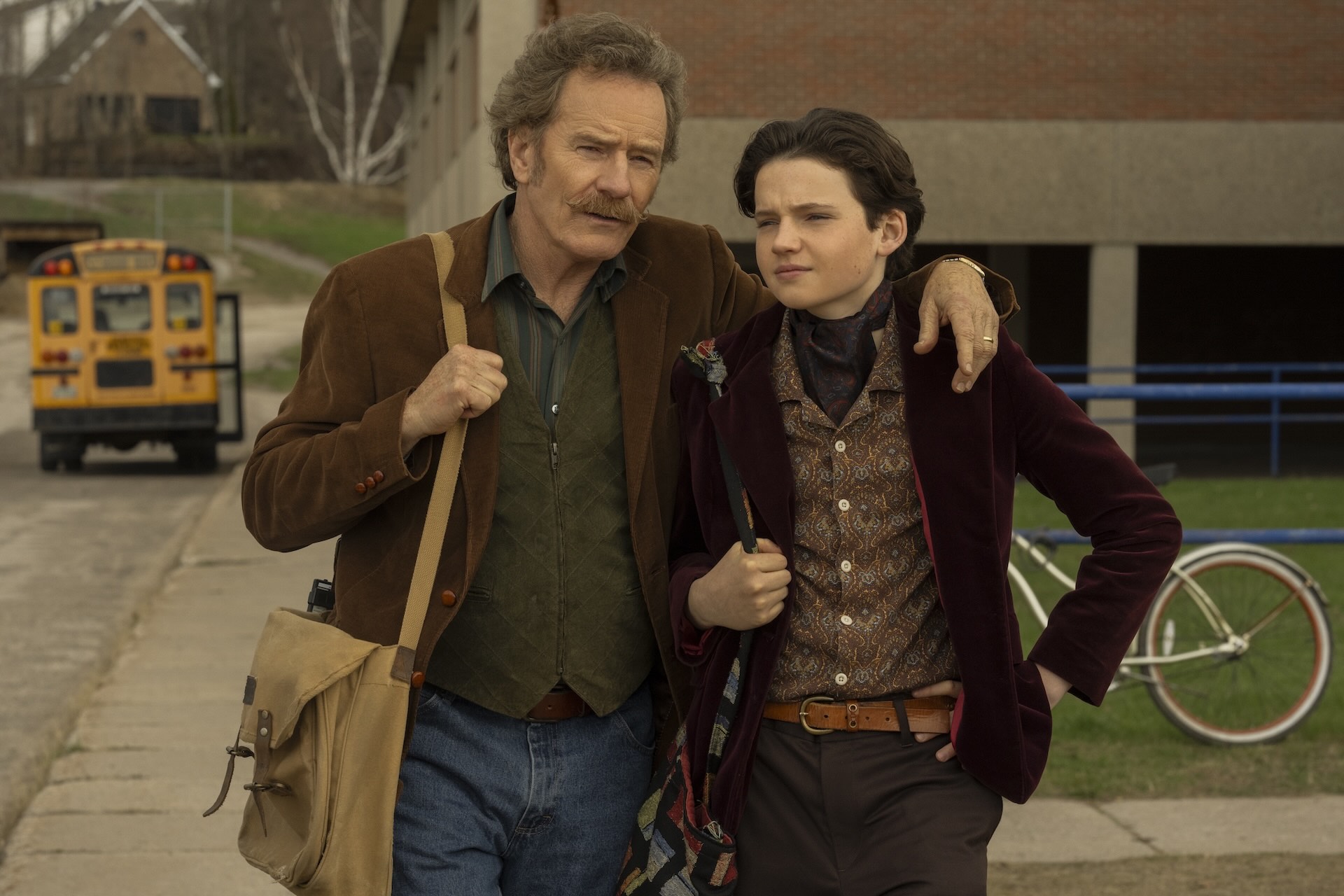 Man and teenager walking on sidewalk; man with mustache and satchel has arm around teen. Yellow bus and bicycle in background.