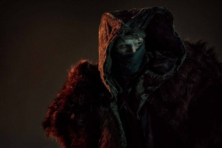 Mysterious figure in a dark hood and fur cloak gazes intensely to the side, face partially obscured.
