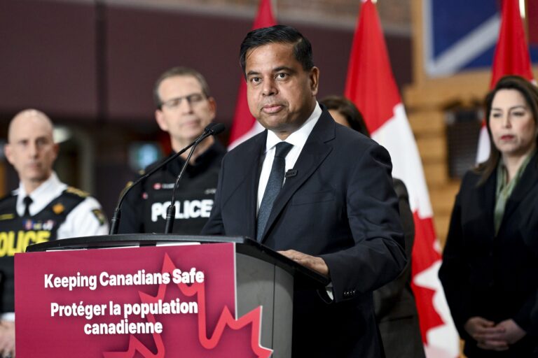 A press conference shot of Gary Anandasangaree at a podium bearing a sign reading, "Keeping Canadians Safe." He is flanked by police officers and others.