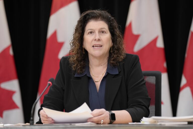 A head-on shot of Karen Hogan seated at a table during a press conference, with Canadian flags on stands in the background.