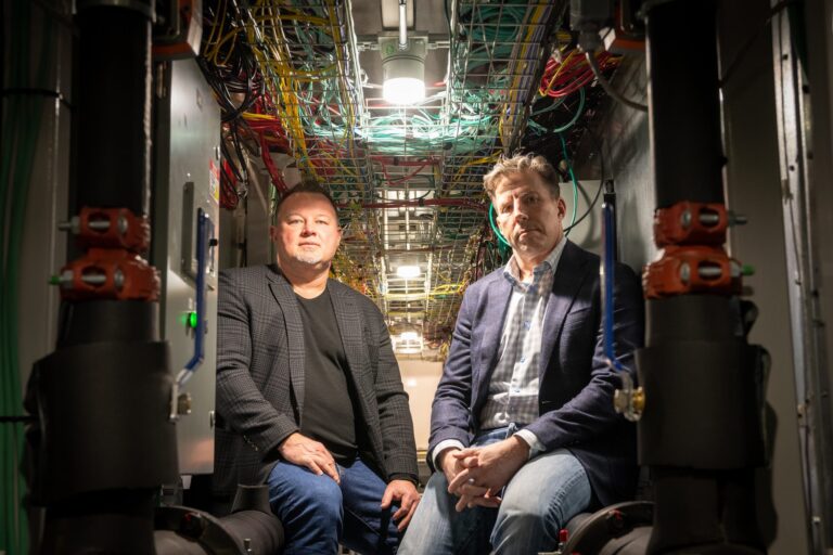 Two men wearing suit jackets and jeans sit in a dark room, facing the camera. They are surrounded by blue, yellow and red wires.