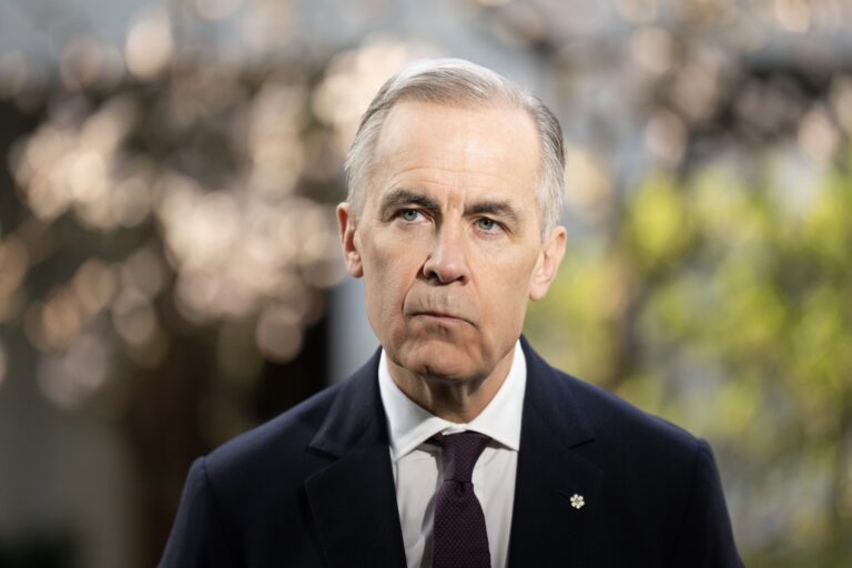 Prime Minister Mark Carney wears a dark suit and tie, with a serious facial expression.