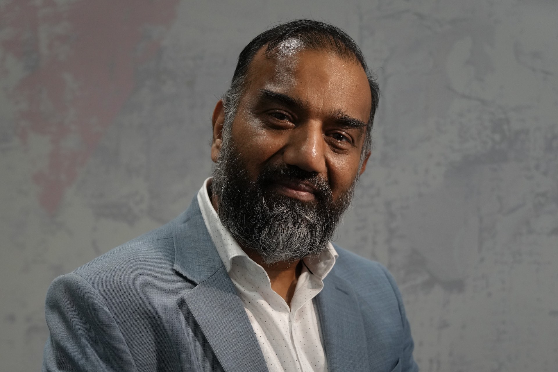 A portrait of Manav Gupta. He's wearing a grey suit with a white, open-necked shirt.