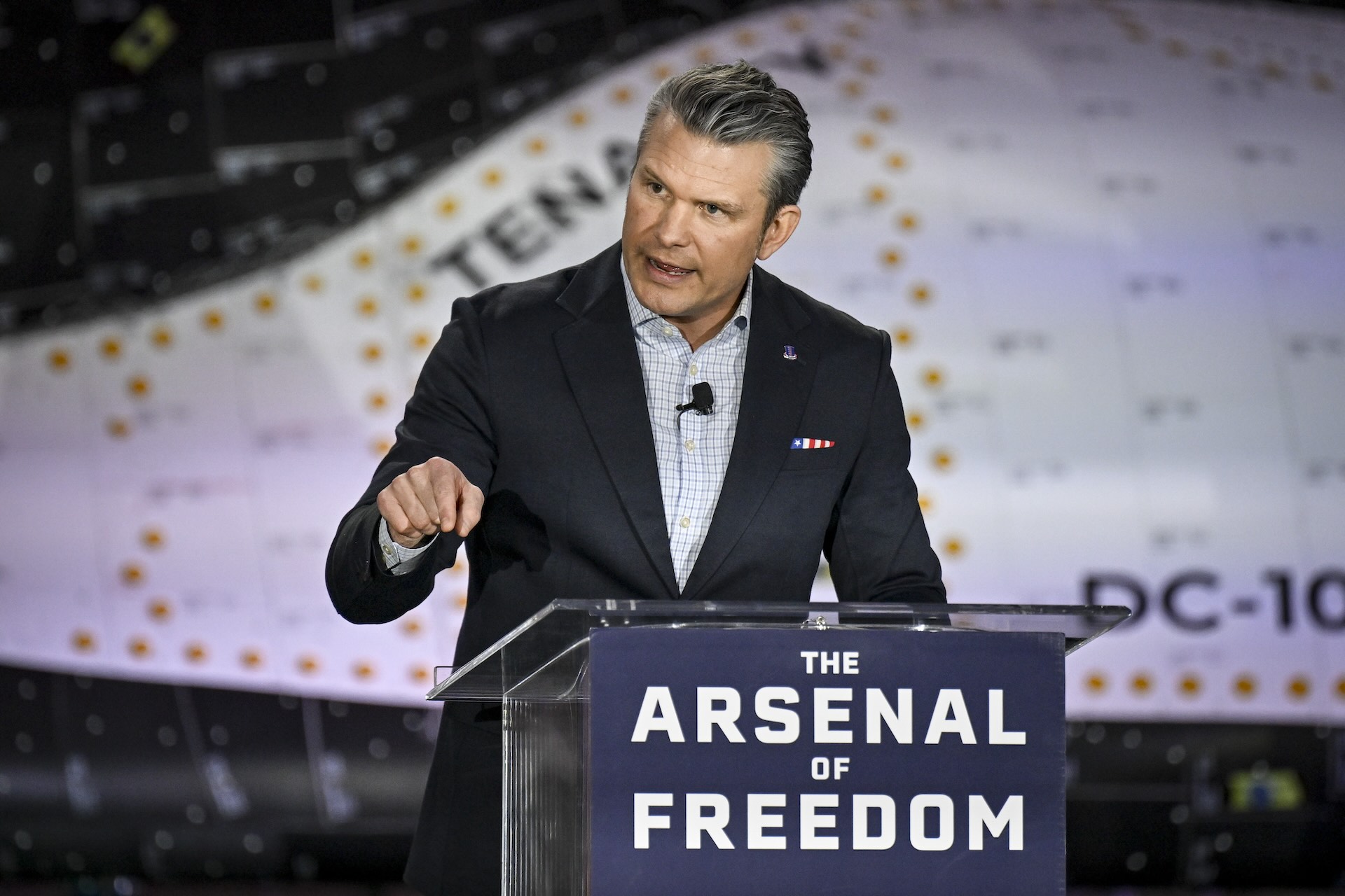 Pete Hegseth speaks at a podium that has a sign reading "The Arsenal of Freedom" on it. Behind him the frame is filled up by the side panels of the Dreamchaser Tenactiy spaceplane.