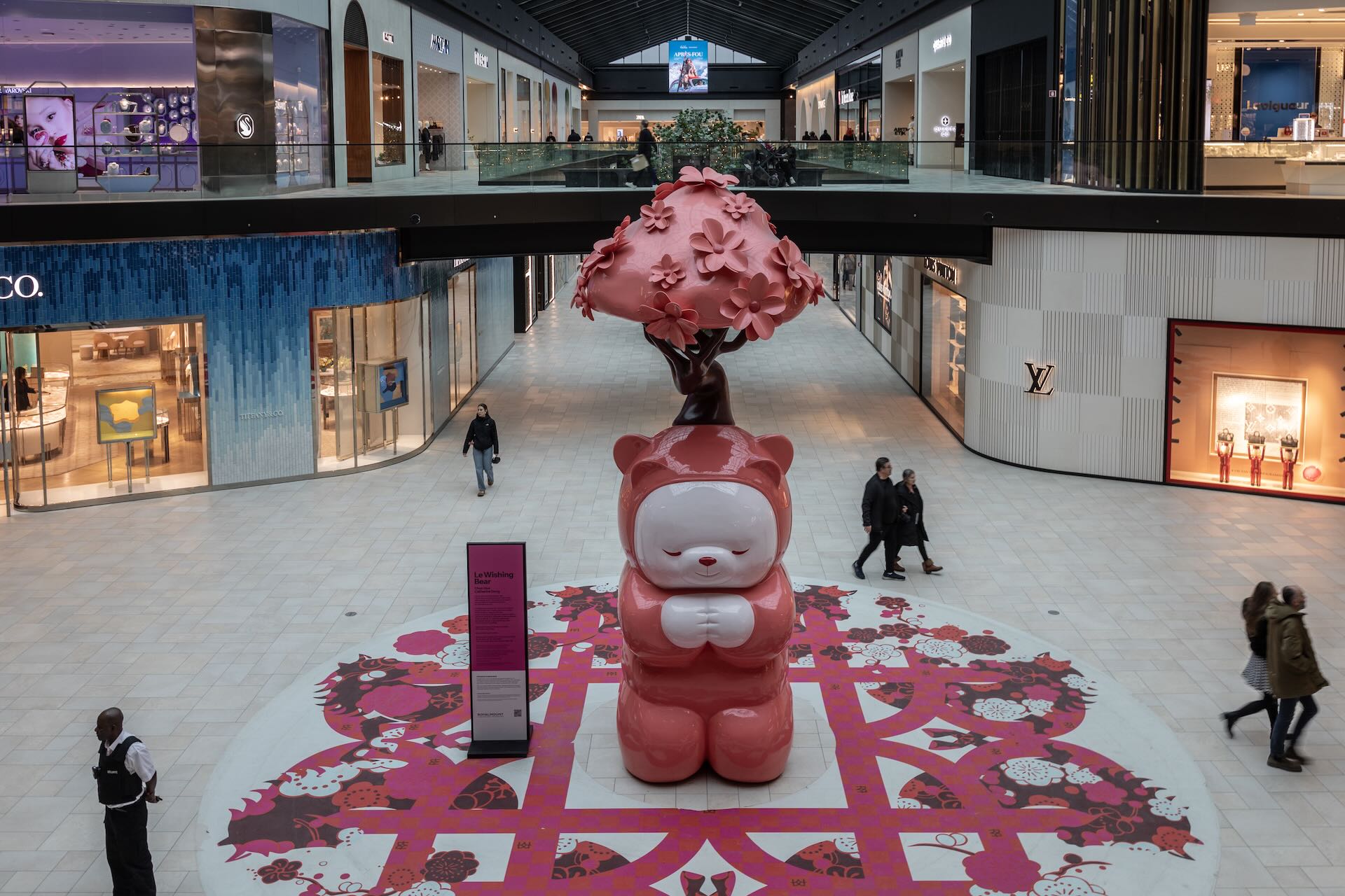 A large, pink fibreglass panda sculpture fills the central court at the Royalmount mall. People are seen walking around the sculpture, with luxury storefronts in the background.