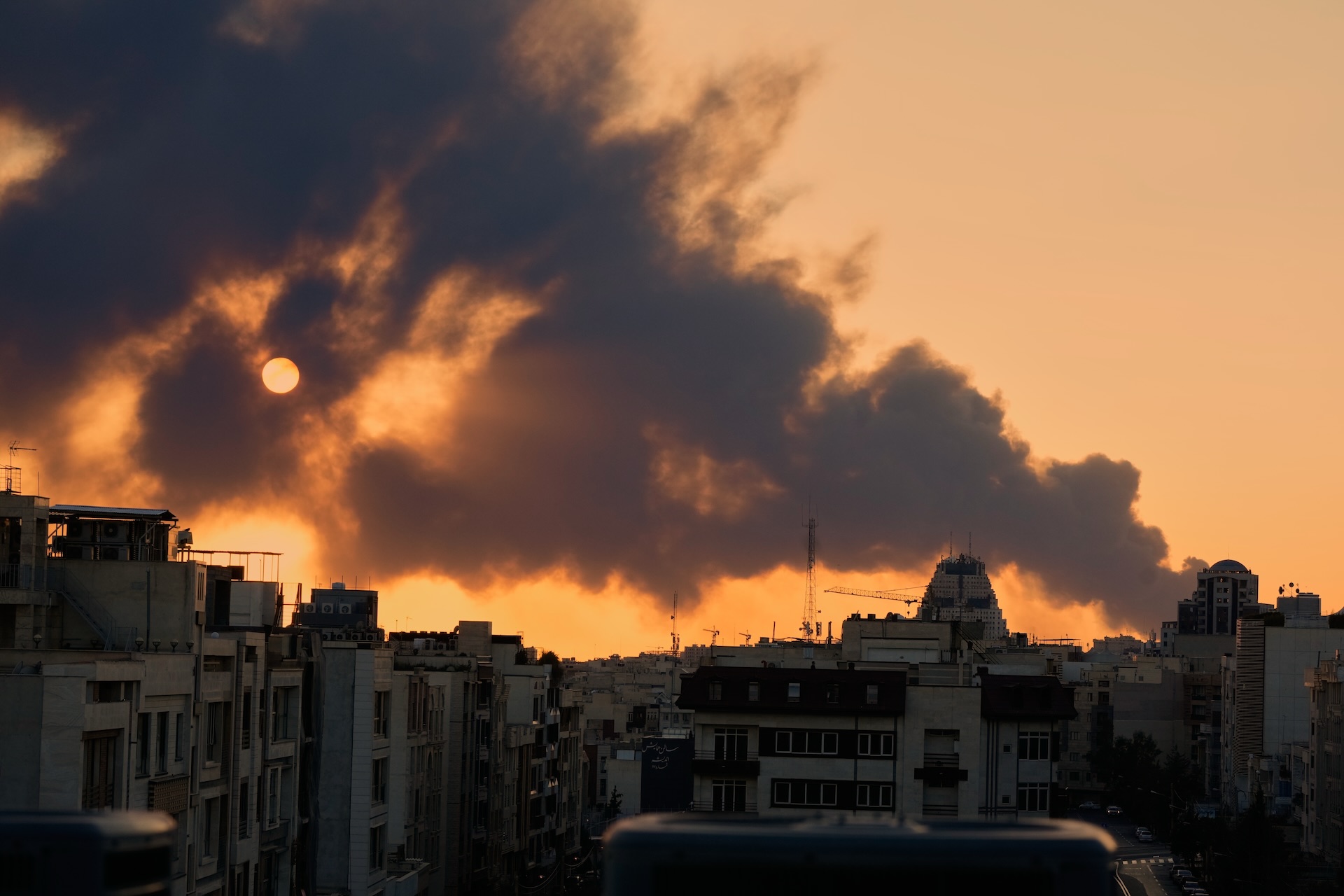 Image of Tehran skyline against a red sky with a plume of smoke billowing from a building, partially obscuring the sun