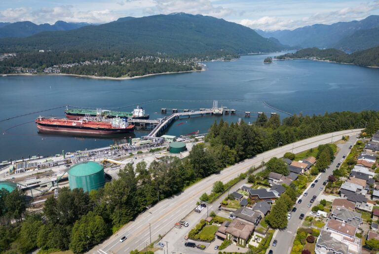 An aerial photo of a petroleum sea terminal, in a landscape of mountains and coastal waterways, with residential homes in the right foreground. There are two tankers docked at the terminal.