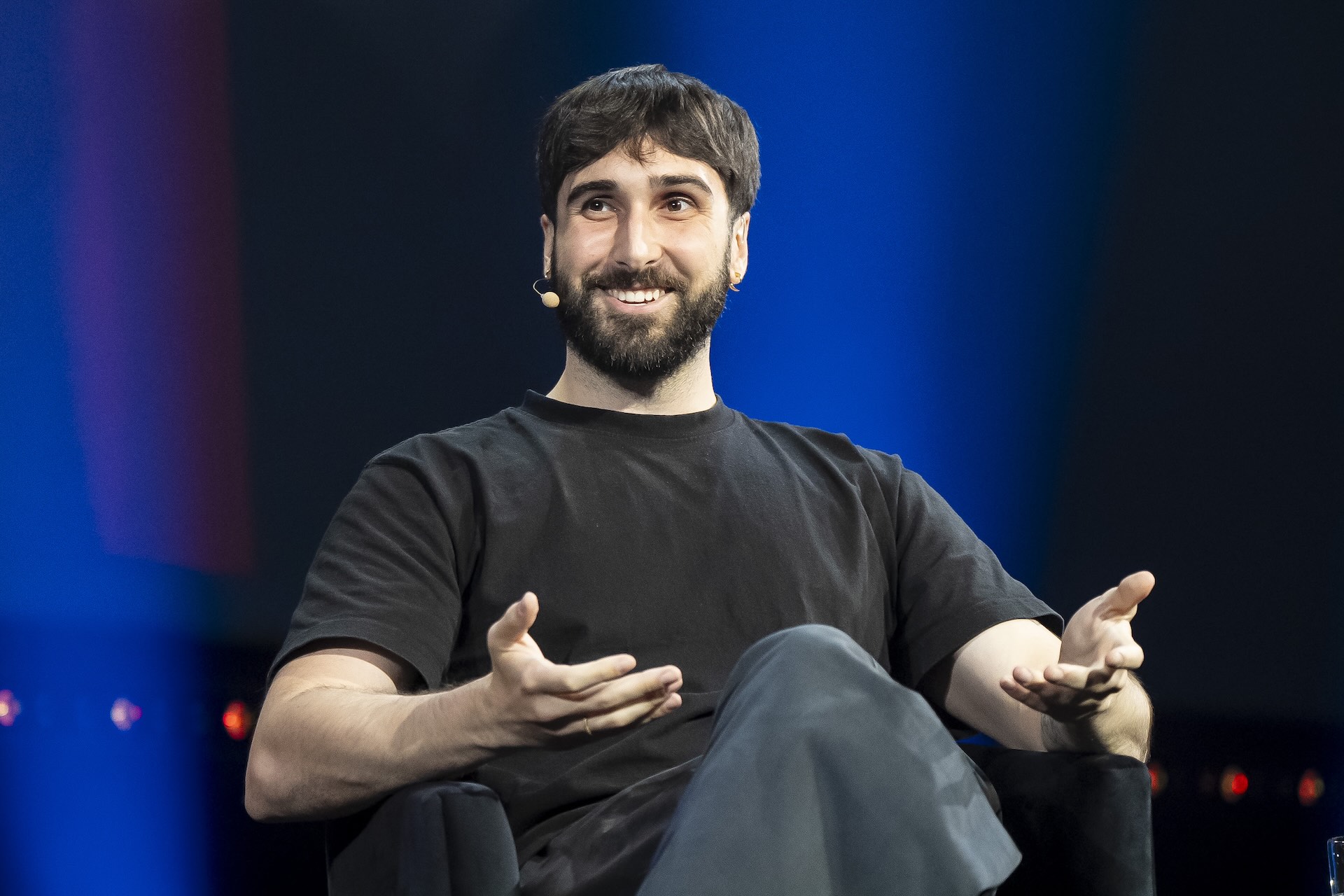 A man wearing a headset microphone sits on a stage. He is speaking and gesturing with his hands. The background is dark blue.