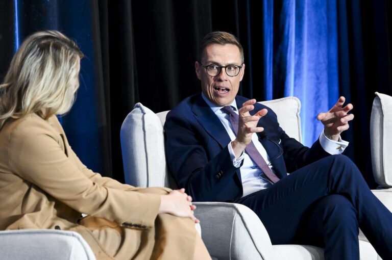 Alexander Stubb in a dark suit and tie gestures while sitting in a white chair on stage and speaking to Mélanie Joly, sitting next to him in another chair and looking away from the camera toward him.