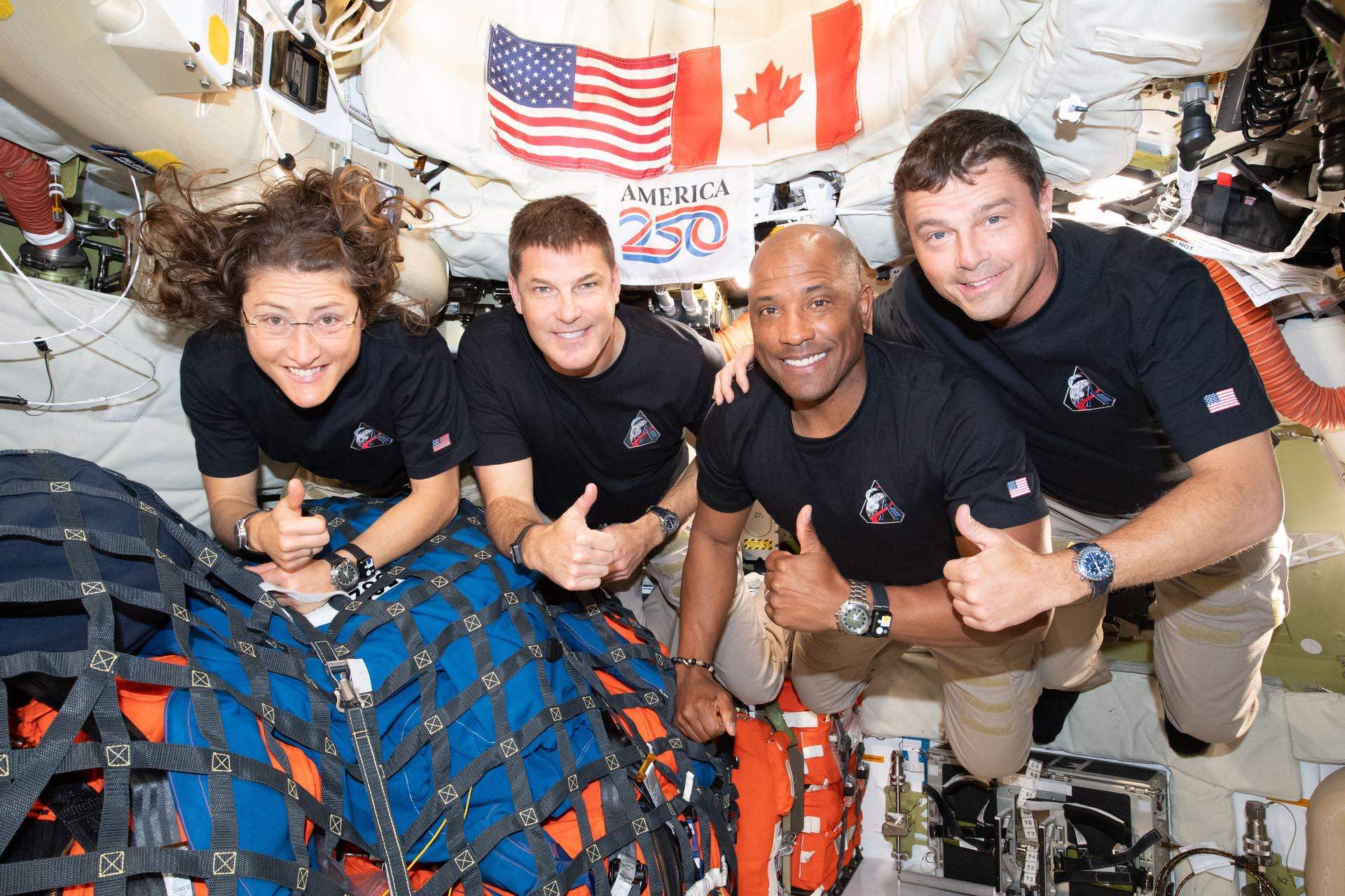A group shot of the Artemis II crew inside their crowded capsule, with Canadian and American flags and an "America 250" flag in the background. They're smiling and giving the thumbs-up. There is gear tied down under netting to the lower left of the frame.