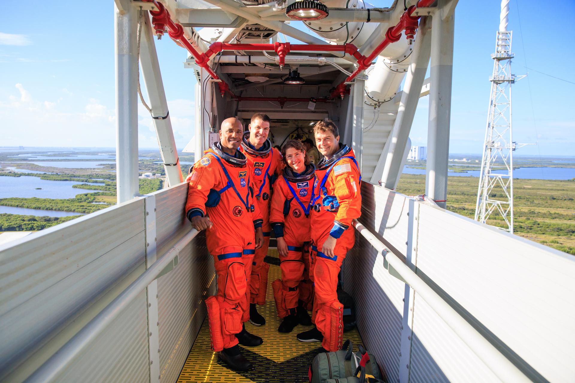 Four astronauts in orange space suits (no helmets) stand on a launch-pad walkway high above ground level, posing and smiling for the camera. Green fields and lakes under blue skies can be seen in the background.