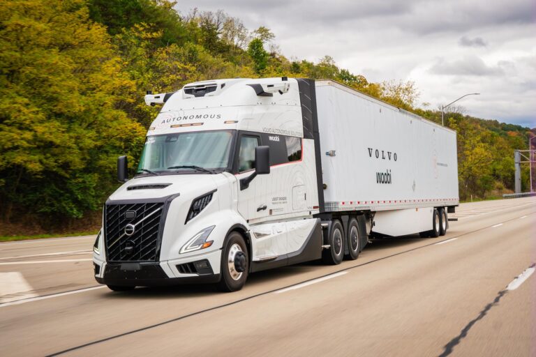 A photo of a white, tandem-axle truck pulling a semi trailer bearing the Volvo and Waabi logos. The truck appears to be on a major highway.