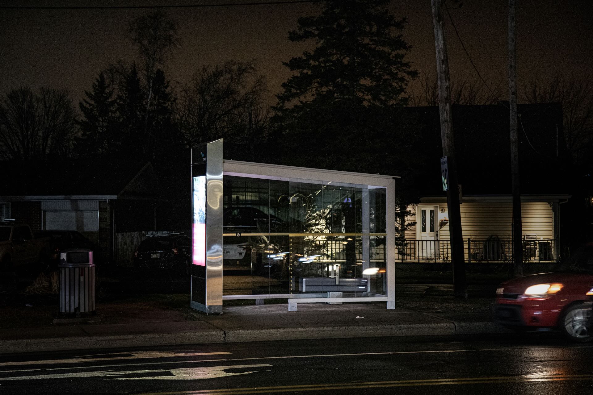 A dimly lit bus stop at night, with a glowing ad panel, stands on a suburban street. A red car passes by, reflecting on the wet road.