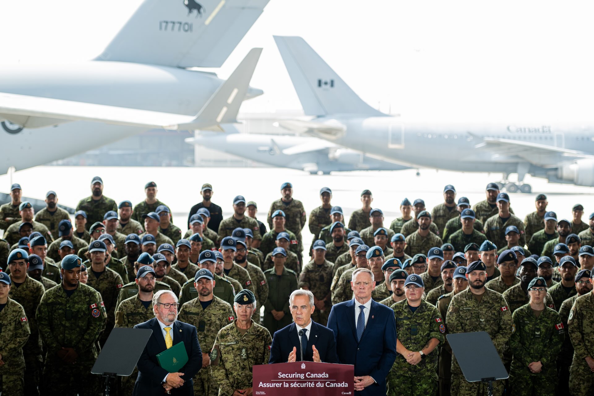 Mark Carney stands at a podium, speaking. Behind him are dozens of people wearing CAF uniforms. In the background, two planes are visible.