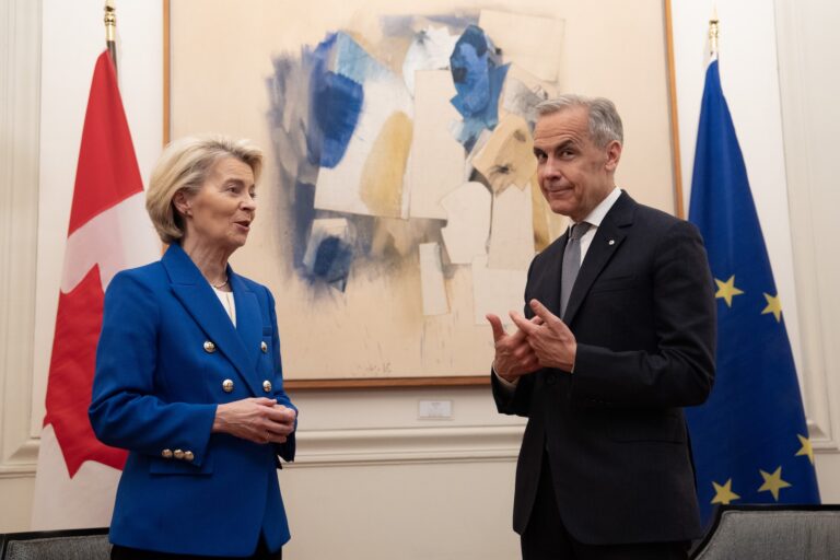 A shot of Mark Carney and Ursula von der Leyen in front of an abstract painting with Canadian and EU flags on either side of it. Von der Leyen is speaking, and Carney appears to be shrugging at the camera with a sly smile on his face.