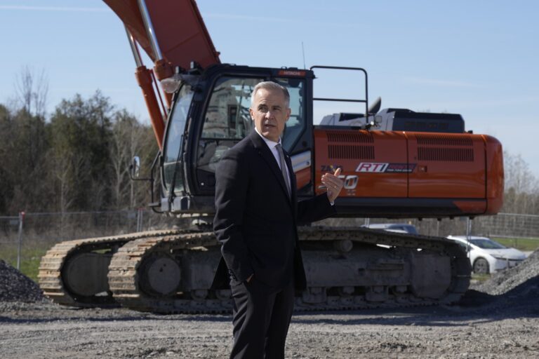 A shot of Mark Carney standing in a dark suit in front of a large, red excavator on a construction site. Carney is looking behind him, off-camera.