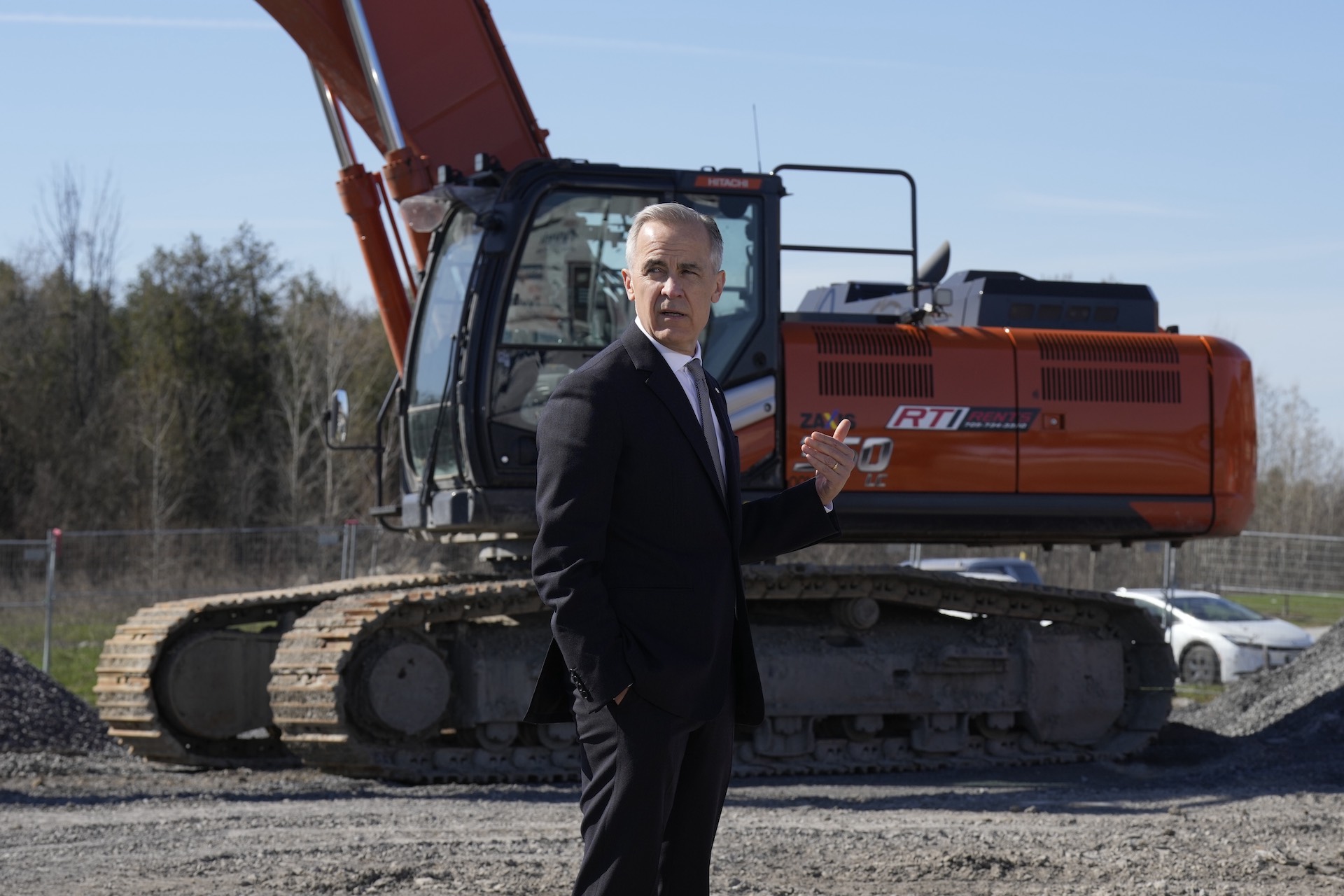 A shot of Mark Carney standing in a dark suit in front of a large, red excavator on a construction site. Carney is looking behind him, off-camera.