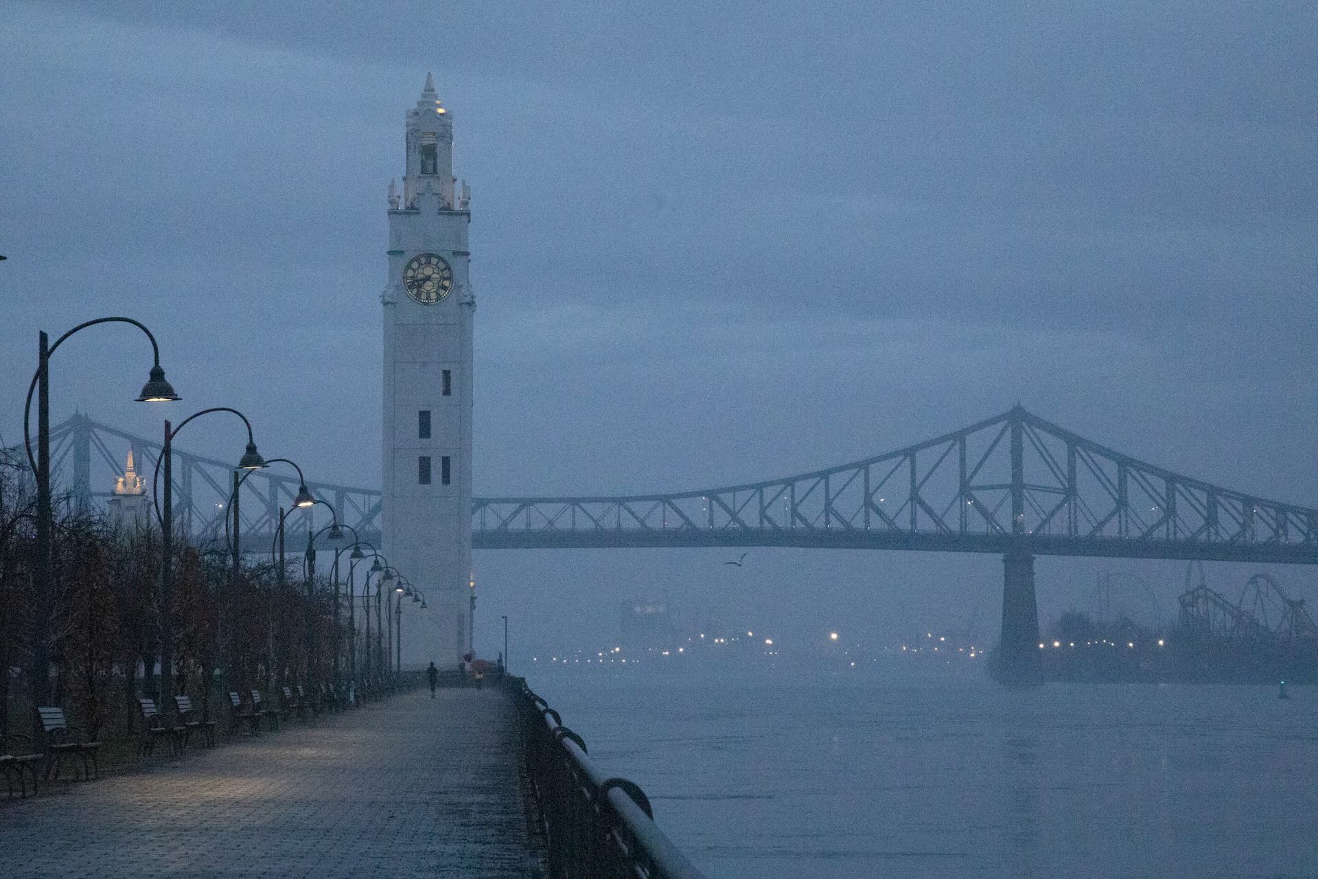 A foggy morning scene shows a riverside path with lampposts leading to a tall clock tower, and a bridge in the background, evoking a serene, misty ambiance.